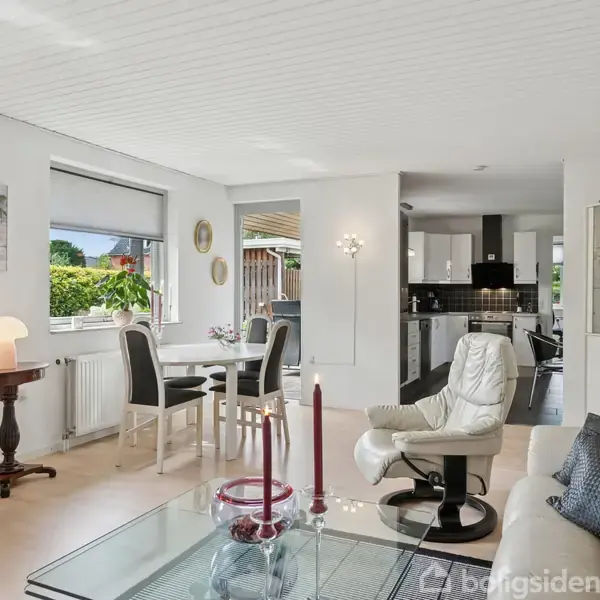 Stue med moderne møbler: To lænestole og en sofa omkranser et glasbord med røde lys. I baggrunden ses en spisebordgruppe ved et vindue. Til højre er et åbent køkken.