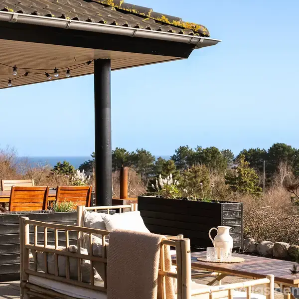 Terrasse med træmøbler og dækket sofa under halvtag. Sofabord med kande og krus. Udsigt mod havet og omkringliggende træer i bakkefyldt landskab. Ingen tekst fundet.