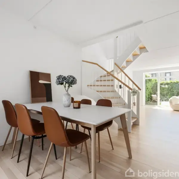 Et lyst spiseområde med et hvidt bord og fire brune stole. Bagved ses en trappe til øverste etage. Store vinduer i baggrunden giver udsigt til en have.