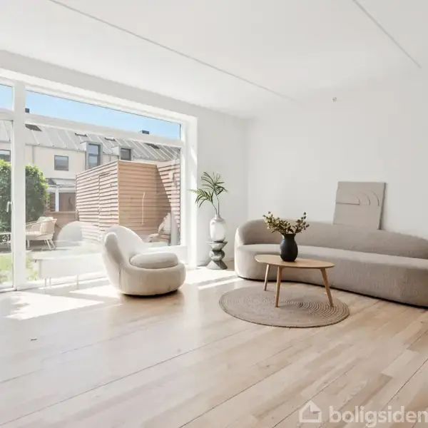 Stue med lys sofa ved væggen, en rund lænestol foran store vinduer, og en plante på gulvet. Udenfor ses en terrasse med træafskærmning og møbler.