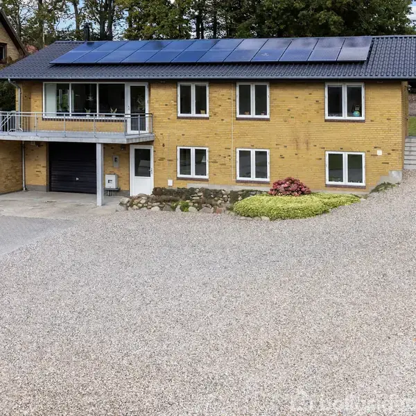 Et gult murstenshus udstyret med solpaneler på taget, beliggende i et stenbelagt gårdsplads. Huset har en balkon over en garage og er omgivet af grønne buske.