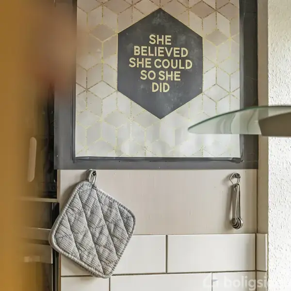 Plakat med teksten "She believed she could so she did" hænger på væggen. Under plakaten er en grå ovnhandske og en ske i et køkkenmiljø.