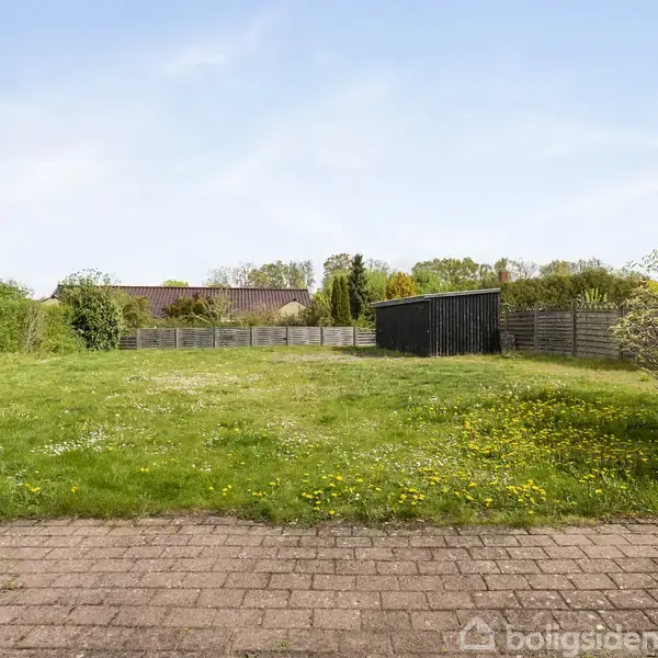 Dør åbner mod en græsplæne med enkelte blomster. I baggrunden ses en mørk træskur og hegn, omgivet af træer og buske under en klar himmel.