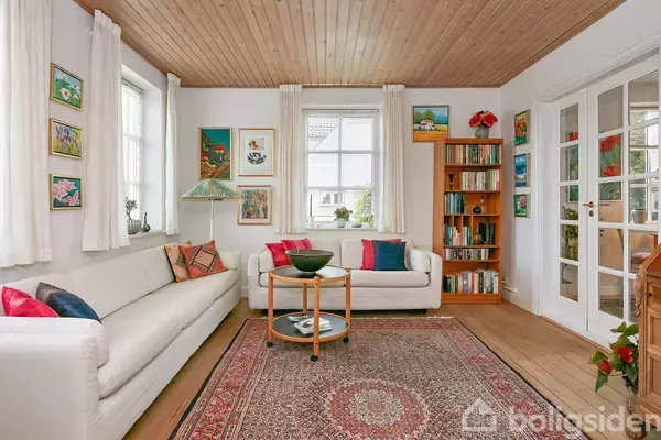 To hvide sofaer står i en stue med malerier på væggene. Et rundt sofabord med skål står på et orientalsk tæppe. En bogreol ved døren fuldender rummet.