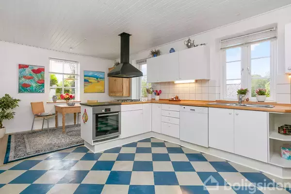 Et lyst køkken med hvid skab og sort emhætte står i et rum med skakternet gulv. Væggene har kunstmalerier, og et spisebord med stole ses ved et vindue.