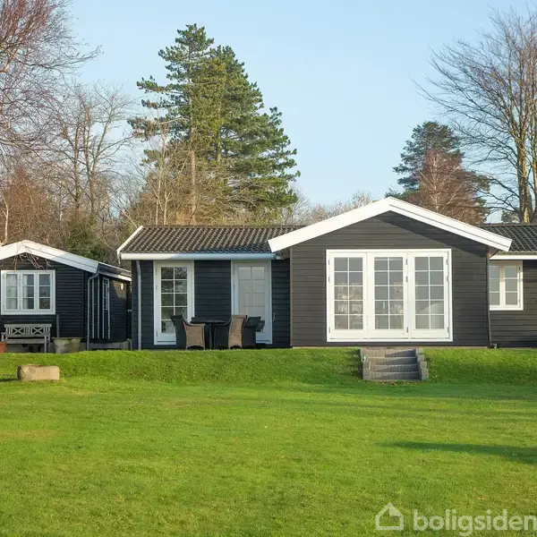 En sortmalet sommerhus med hvide vinduer står på en velplejet græsplæne omgivet af træer. Huset har store terrassedøre og en mindre udhus til venstre.