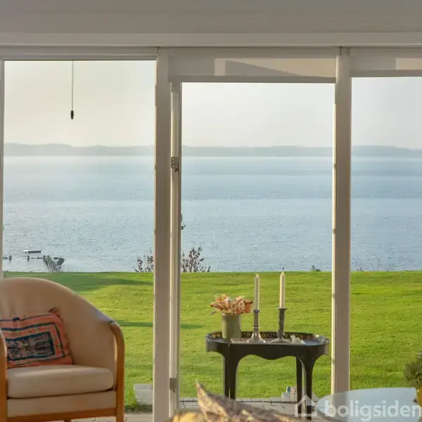 En lænestol står i stuen med udsigt gennem store glasdøre til en grøn græsplæne og en rolig sø, der strækker sig mod horisonten under en klar himmel.