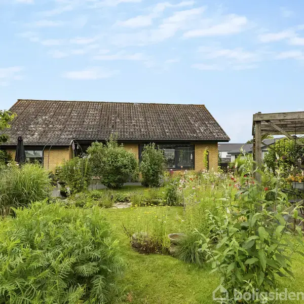 En gul murstensbygning står i en frodig have med grønne planter og buske. Haven har en pergola til højre, der er dækket af klatreplanter. Himlen er blå med enkelte skyer.