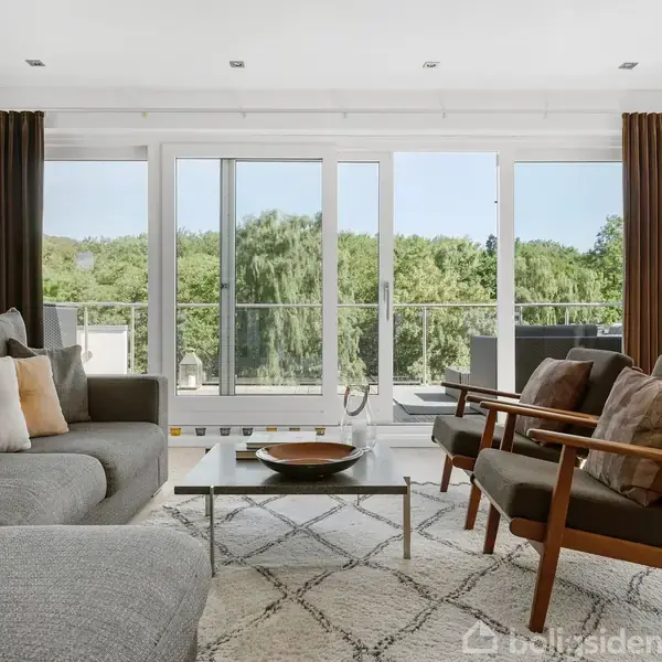 Flere moderne huse står stille på en bakke, omgivet af frodig skov og velplejede haver i et grønt, kuperet boligområde.