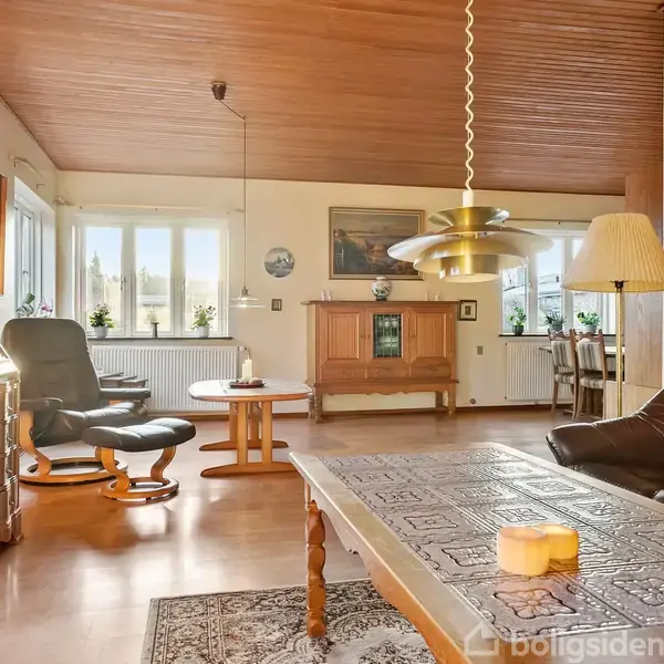 Stue med brune lædermøbler står i et rum med træloft og gulv. Store vinduer slipper lys ind. Et rundt bord med stole ses nær vinduerne. Ingen tekst fundet.