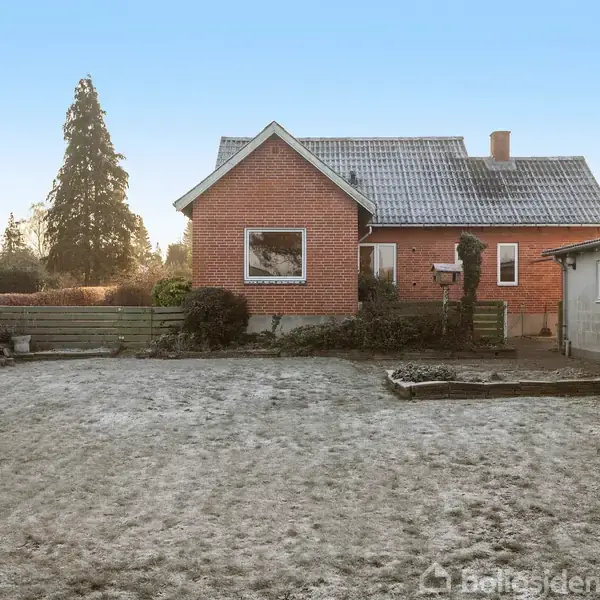 Rødt murstenshus står midt i en frostklædt have med bar jord og omgivende træer. Et tilstødende, hvidt garagebygning er synlig til højre.