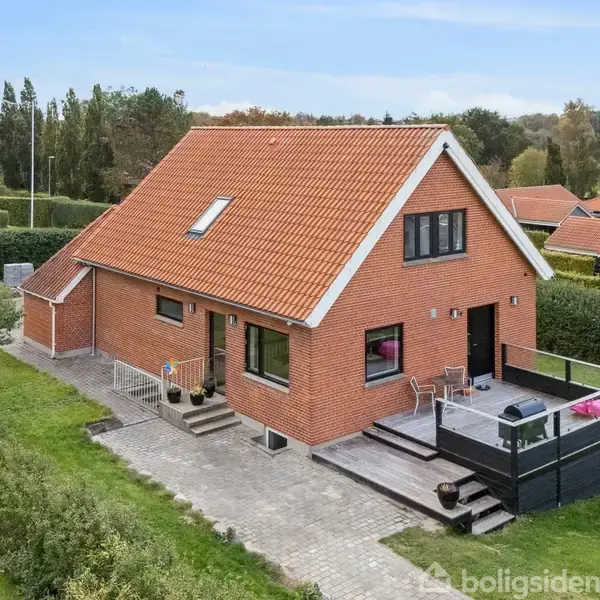 Et rødt murstenshus med rødt tag står på en grøn græsplæne. Foran huset er der en stenterrasse med sorte møbler. Omgivet af hække og naboer i baggrunden.