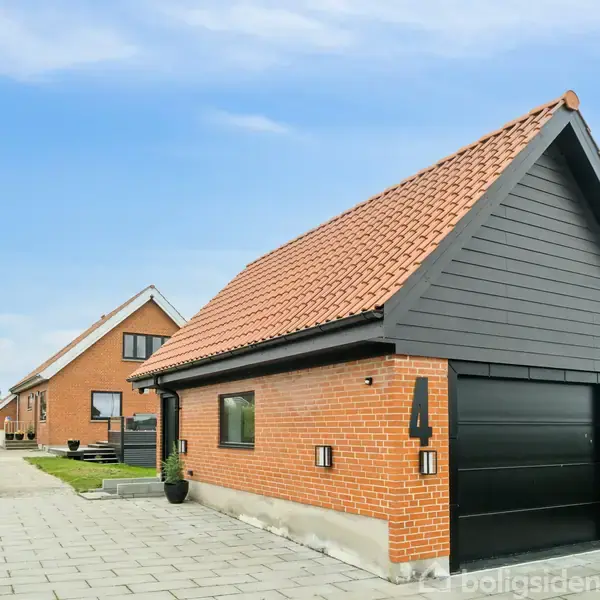 Garagen med rødt tegltag står ved indgangen til en indkørsel foran et murstenshus. Nummeret "4" er synligt på garagens væg. Husene er beliggende i et grønt boligområde.