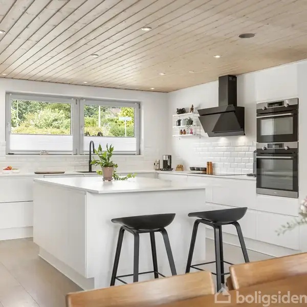 Et moderne køkken med hvidt interiør og et stort vindue. En køkkenø med to sorte barstole er centralt placeret. Blomster og planter tilføjer farve til rummet.