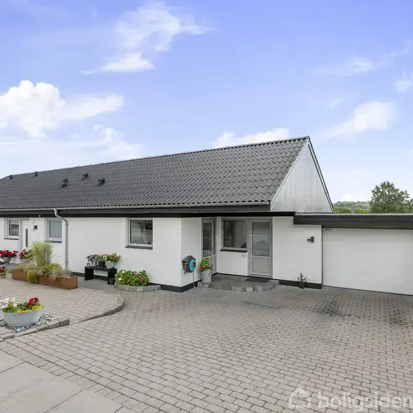 Et hvidt enfamiliehus med sort tag står stille i et boligkvarter. Foran ses en flisebelagt indkørsel med blomsterkrukker og grønne buske langs kanten.