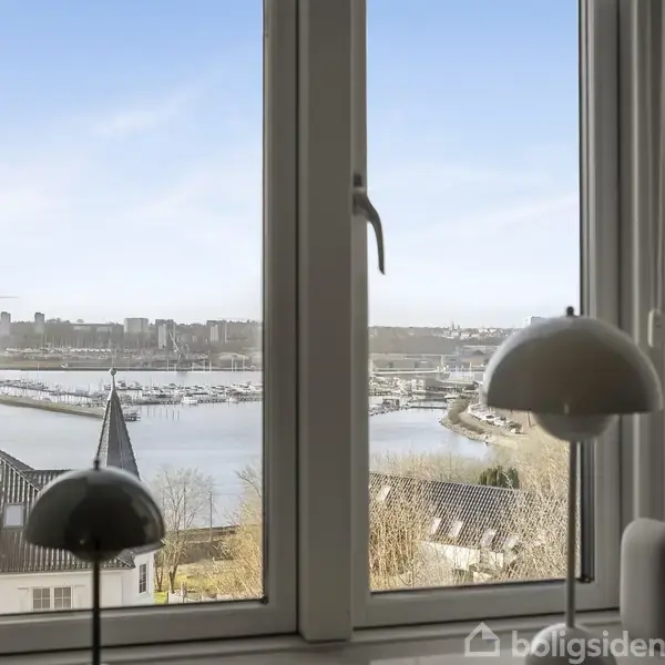 To vinduer viser udsigt til en havn med både og bybygninger i baggrunden. Indendørs lamper placeres nær vindueskarmen, og himlen er klar.