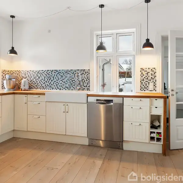 Et moderne køkken med L-formet træbordplade med hvide skabe, en vask, opvaskemaskine og mosaikfliser på væggen. Hængende lamper lyser rummet med lyse trægulve op. Ingen tekst fundet.