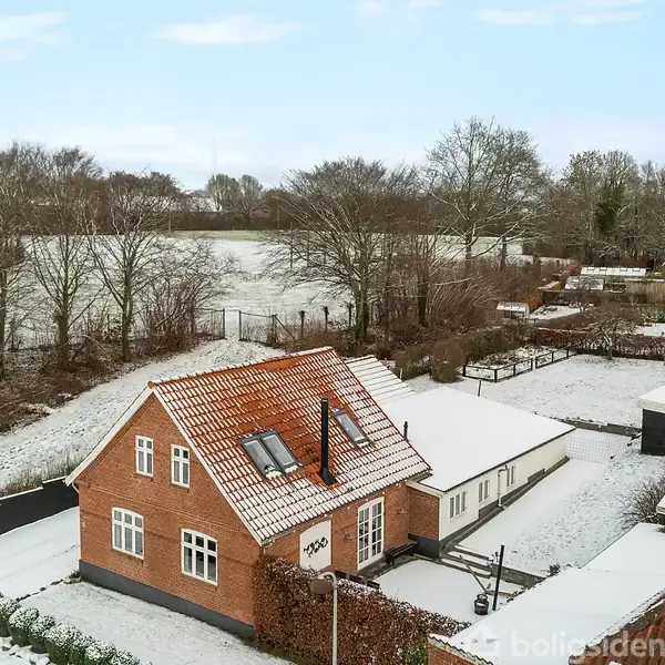 Et rødt murstenshus med rødt tag står stille omgivet af snedækkede marker og træer. Der er små udhuse ved siden af huset, og haven er dækket af sne.