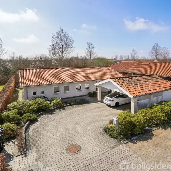 Huset har rødt tegltag og er omgivet af en brolagt indkørsel med grønne buske. En bil er parkeret under en overdækket carport. Skovområde i baggrunden.