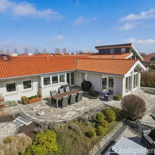 Et hvidt hus med rødt tag står i en anlagt have. En stenbelagt terrasse med havemøbler ligger foran huset. Omkring er velplejede buske og træer under en blå himmel.
