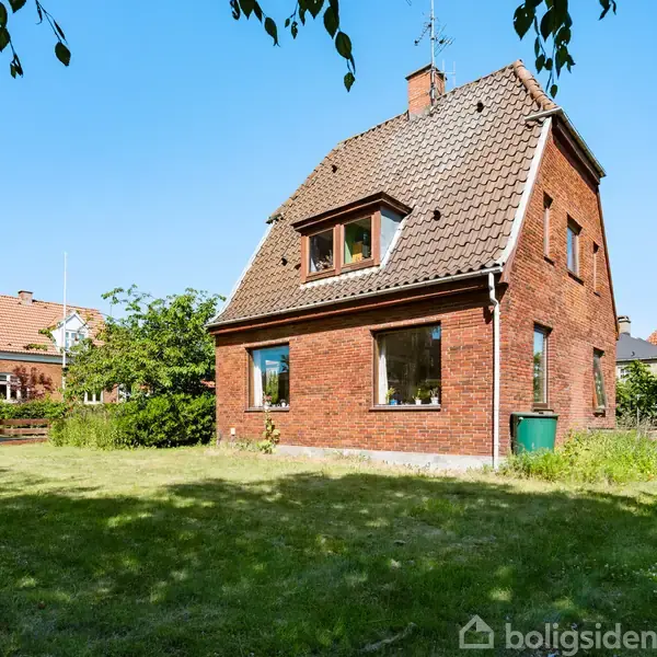 Rødstenshus står stille i en grøn have med blå himmel. Omkring huset er der græsplæne og buske, mens andre huse kan ses i baggrunden.