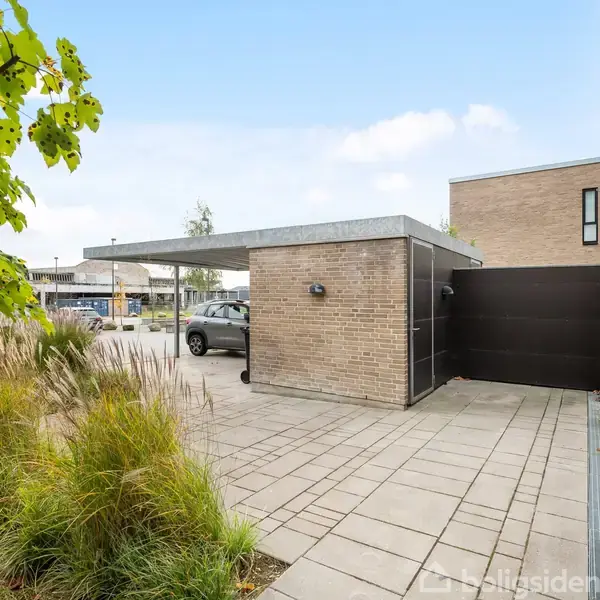 En bil er parkeret under en overdækket carport ved et murstenshus. Græsser og buske omgiver flisebelægningen i forgrunden, mens en lys, solrig himmel danner baggrunden.