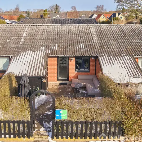 Rækkehus med røde mursten og tag med sne. I forhaven er der møbler og en sort parasol. Et skilt i forhaven viser "Til salg - Nybolig" omgivet af hække.