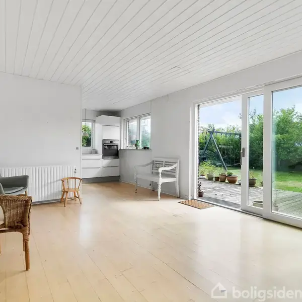 Lys stue med trægulv og hvide vægge, indeholder to stole og et bord. Store vinduer åbner mod en terrasse og frodig have udenfor.