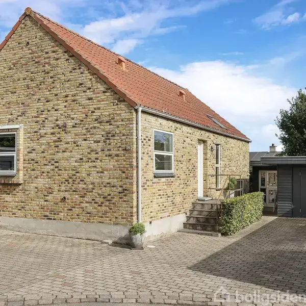 Rødstenshus med rødt tag står stille på en brostensbelagt gårdsplads. Til højre ses en grå garage og grønne buske. Himlen er blå med lette skyer.