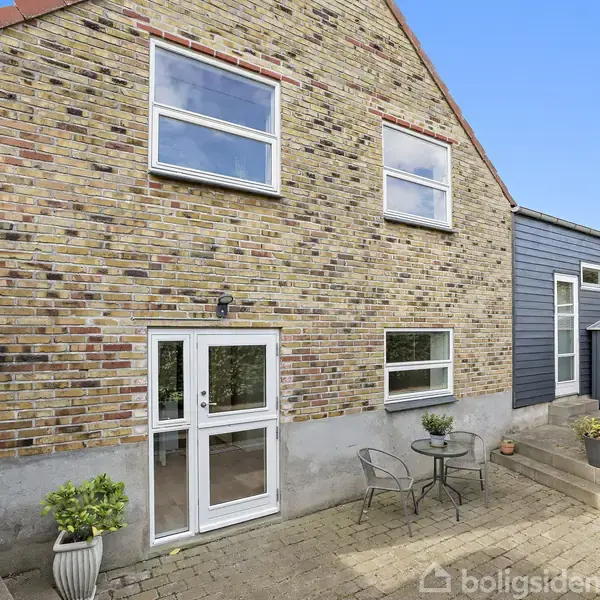 To-etagers murstenshus med store vinduer og en havedør, står i en flisebelagt gårdhave med et bord og to stole omgivet af blomsterkrukker.