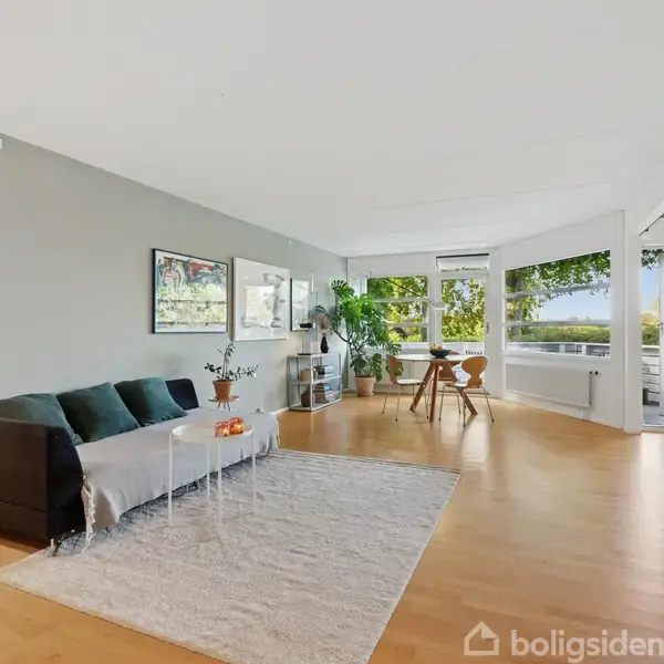 Stue med sort sofa ved siden af et lille bord på et lyst tæppe, udsigt til træer gennem store vinduer omkranser rummet, der er møbleret med spisestuebord og stueplante.