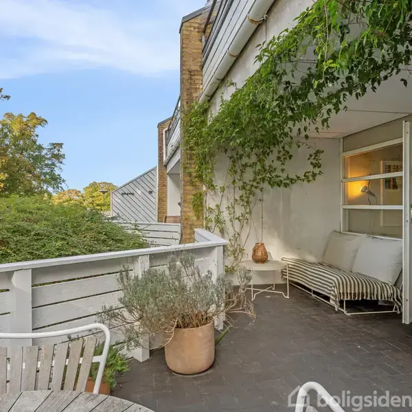 Terrasse med træstole ved rundt bord, omkranset af hvidt hegn. Langs væggen vokser grønne klatreplanter. En åben dør leder ind til et minimalistisk indrettet rum.
