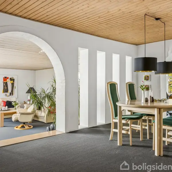 Bord og stole står i spisestue med mørkt tæppe, lys træloft og moderne lamper. Bueformede arkader skiller spiseområde og dagligstue med hvide sofaer og dekorationer i et lyst og rummeligt miljø.
