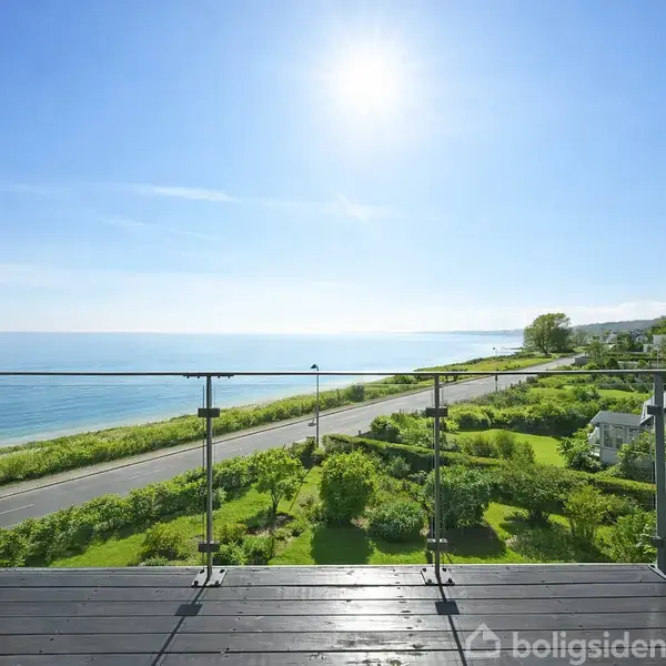 Døre står åbent mod en balkon, der tilbyder udsigt over en kystvej, grønne buske og havet under en klar blå himmel og strålende sol.