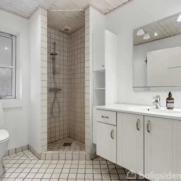 Toilet i moderne badeværelse. Bruser i hjørnet, fliser på gulvet, hvidt vaskeskab med spejl. Enkel indretning, neutralt farveskema. Flasken på bordet har etiketten vendt væk.