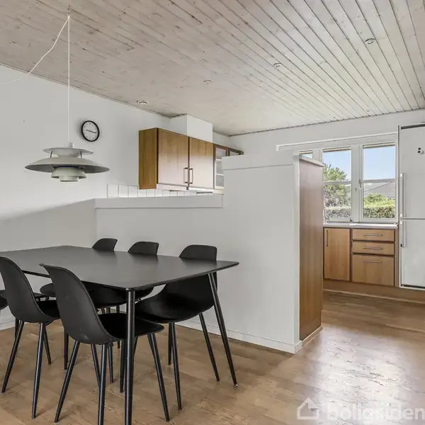Spisebord med sorte stole står i en spisestue. En pendellampe hænger over bordet. Køkken med træskabe og et vindue ses i baggrunden. Vægur hænger på væggen.