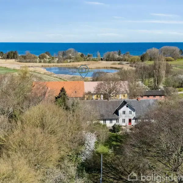 En gård med røde tage ligger omgivet af træer og marker. I baggrunden ses en dam og havet. Klar blå himmel over det åbne landskab.