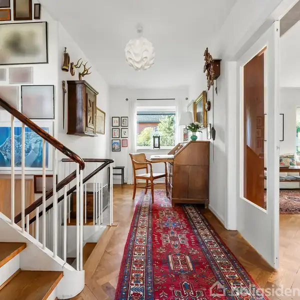 Trappe med trætrin fører opad; væggen omkring er dekoreret med kunstværker. I gangen ligger en rød orientalsk løber. I baggrunden ses et skrivebord med vindue og stole omkring.