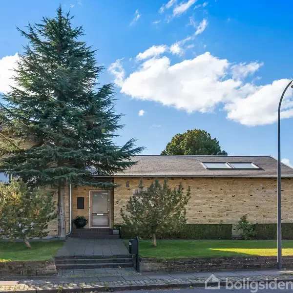 Hus står stille på en parcel med grøn græsplæne og træer omgivet af en fortov og en vej. Følger minimalistisk arkitektur med et skråt tag, ovenlys og murstensfacade.