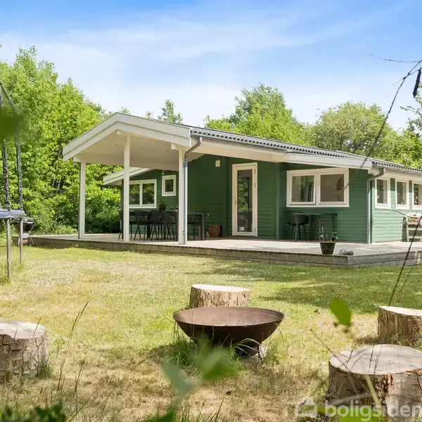 Grønt sommerhus står stille i en frodig have med træstubbe omkring et ildsted og en trampolin på græsset. Skoven omgiver stedet, hvilket skaber en rolig atmosfære.