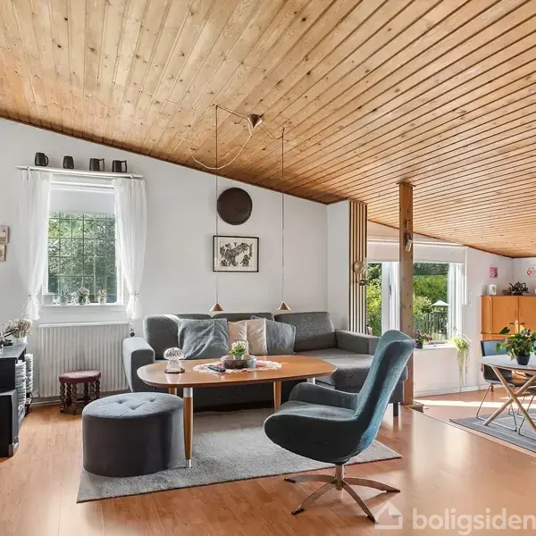 Stue med moderne møbler inkluderer en grå sofa, runde borde og en blå lænestol. Store vinduer lukker naturligt lys ind, mens planter og kunst skaber en hyggelig atmosfære.