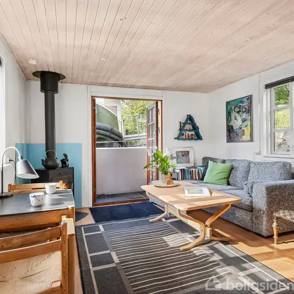Et hyggeligt rum med en grå sofa, sofabord, brændeovn, og skrivebord. Døren er åben mod en terrasse, omgivet af grønne områder og dekoreret med kunstværker på væggene.