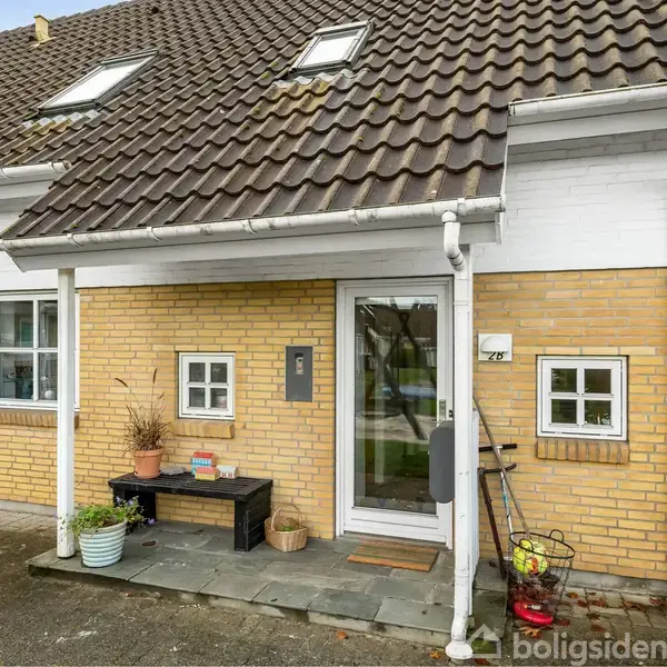 Rækkehus med skråt tag og tre kviste; indgang med glasdør, bænk, planter og cykel ved siden af. Gul murstensfacade og vinduer med hvide karme.