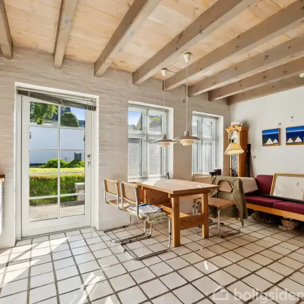 Et køkken-alrum med træmøbler og fliser på gulvet. Et bord med stole står centralt, en dør og vinduer til venstre giver udsigt til en grøn have. Væghængte lamper og malerier dekorerer rummet.