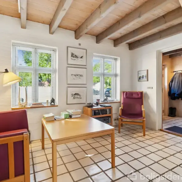 En stue med lyse trægulve og loftsbjælker. En rød sofa og lænestol står ved et træbord. Ophængte billeder pryder væggene. Hallen er synlig gennem en åbning.