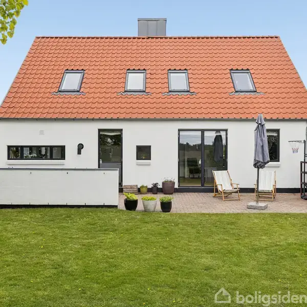 Hus med hvid facade og rødt tegltag, har fire tagvinduer. Foran ses en terrasse med parasol og stole. Omkranset af en grøn græsplæne med træer i baggrunden.