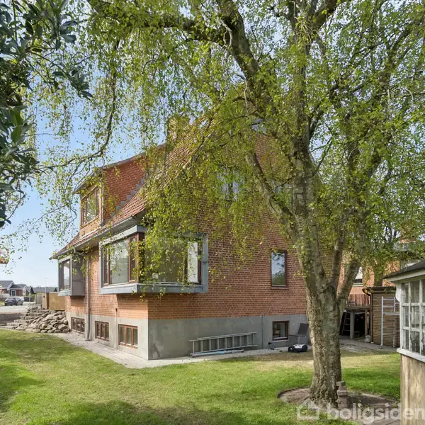 Rødt murstenshus står i en have med græsplæne. Et stort træ med grønne blade hænger forgrenet over huset. På højre side ses en træskur.