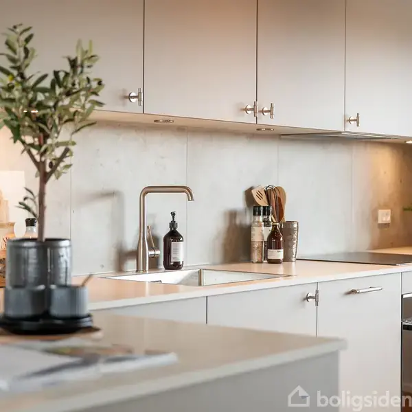 Moderne køkken med hvide skabe og stålgreb. Planterne på bordet og diverse køkkenredskaber ved vasken skaber en hyggelig atmosfære. Marmorvase tilføjer elegance til rummet.