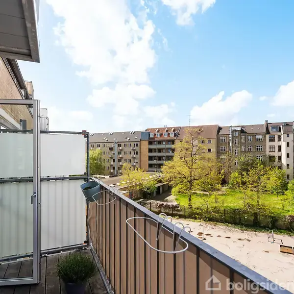 En balkon med glasafskærmning er åben mod en gårdsplads med træer og buske. Bygninger omgiver området under en blå himmel med skyer.