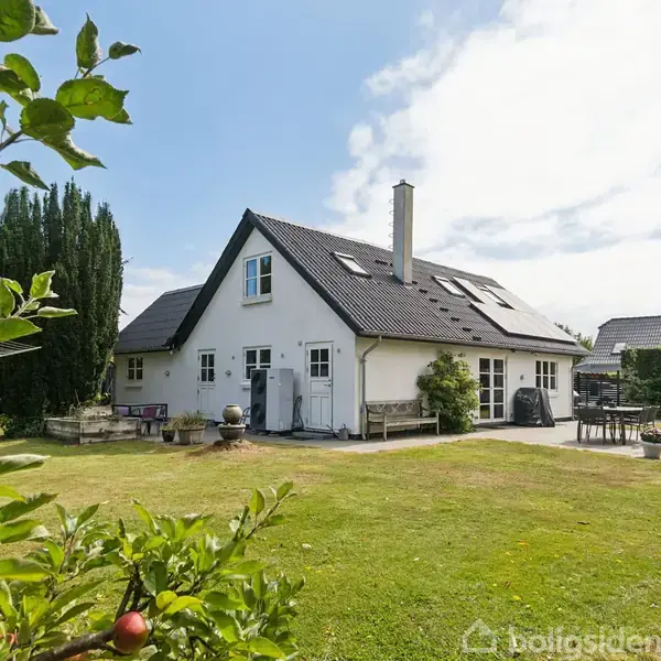 Huset er en hvid to-etagers bygning med skråt tag, omgivet af en grøn have med græsplæne og træer. Et bord og parasol står i haven under en klar blå himmel.